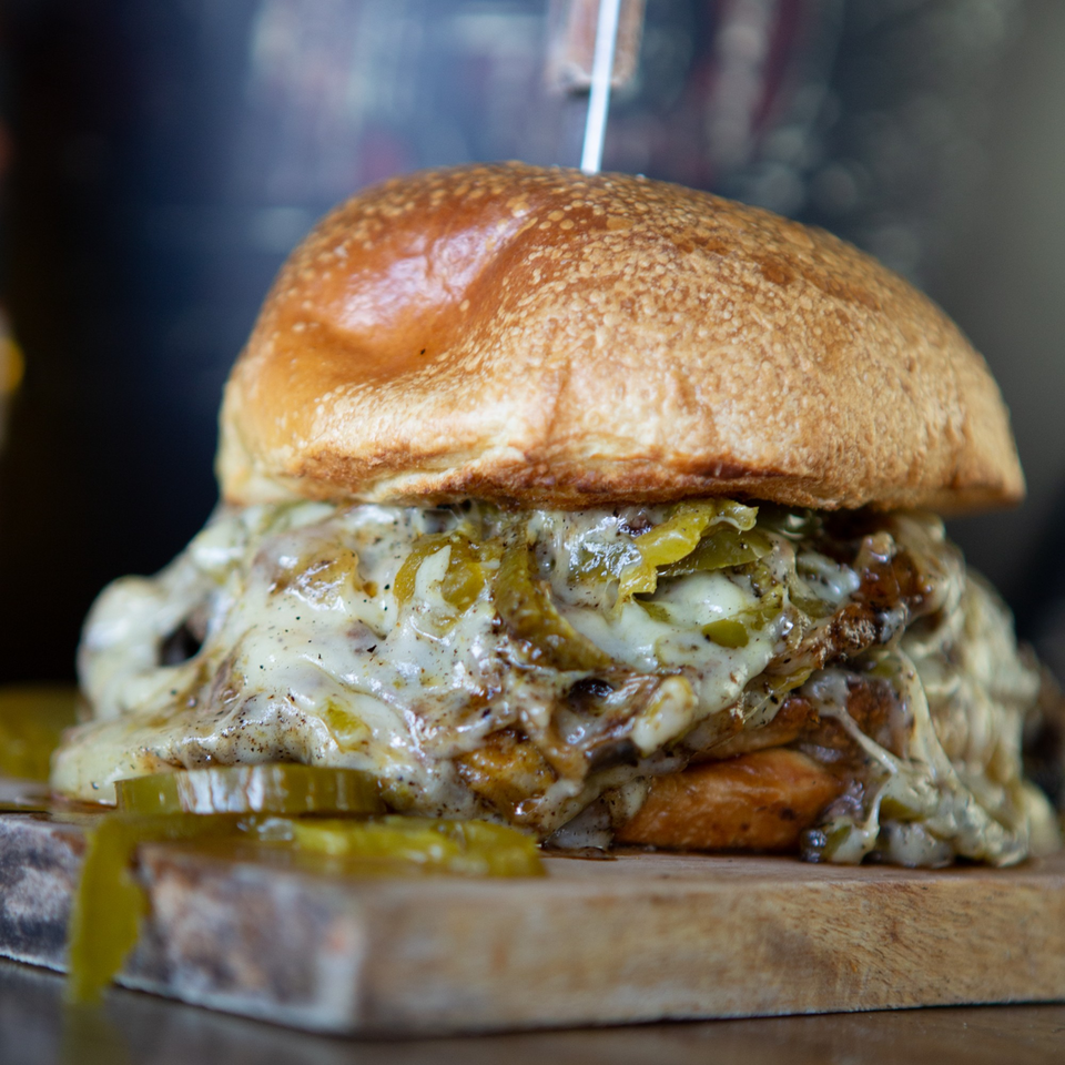 A Blues Burgers Hallandale Beach burger, overflowing with melted cheese and green peppers, on a wooden board.