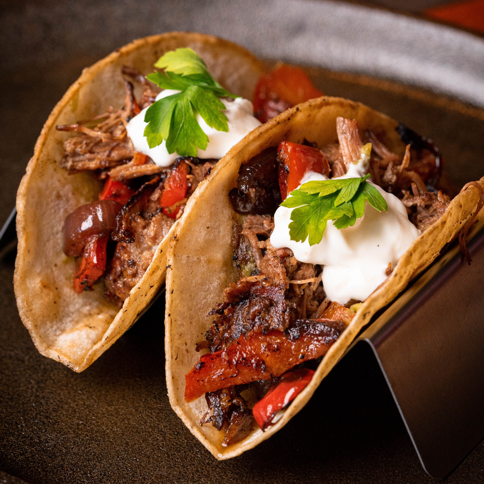 Two beef tacos with red peppers, sour cream, and parsley in a metal holder.